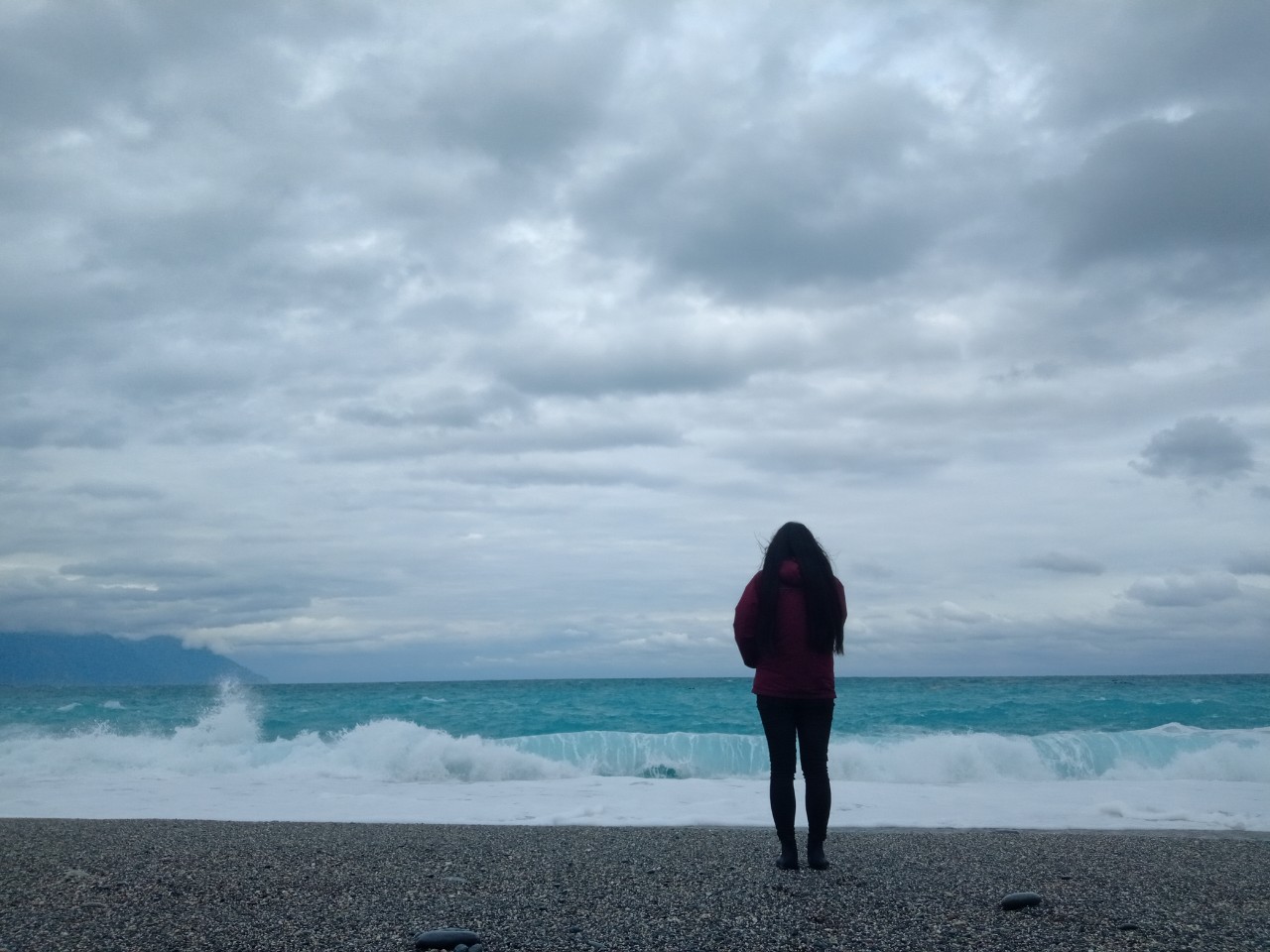 a woman with long hair. her back is to the camera, and she stands on the beach in front of the teal sea.